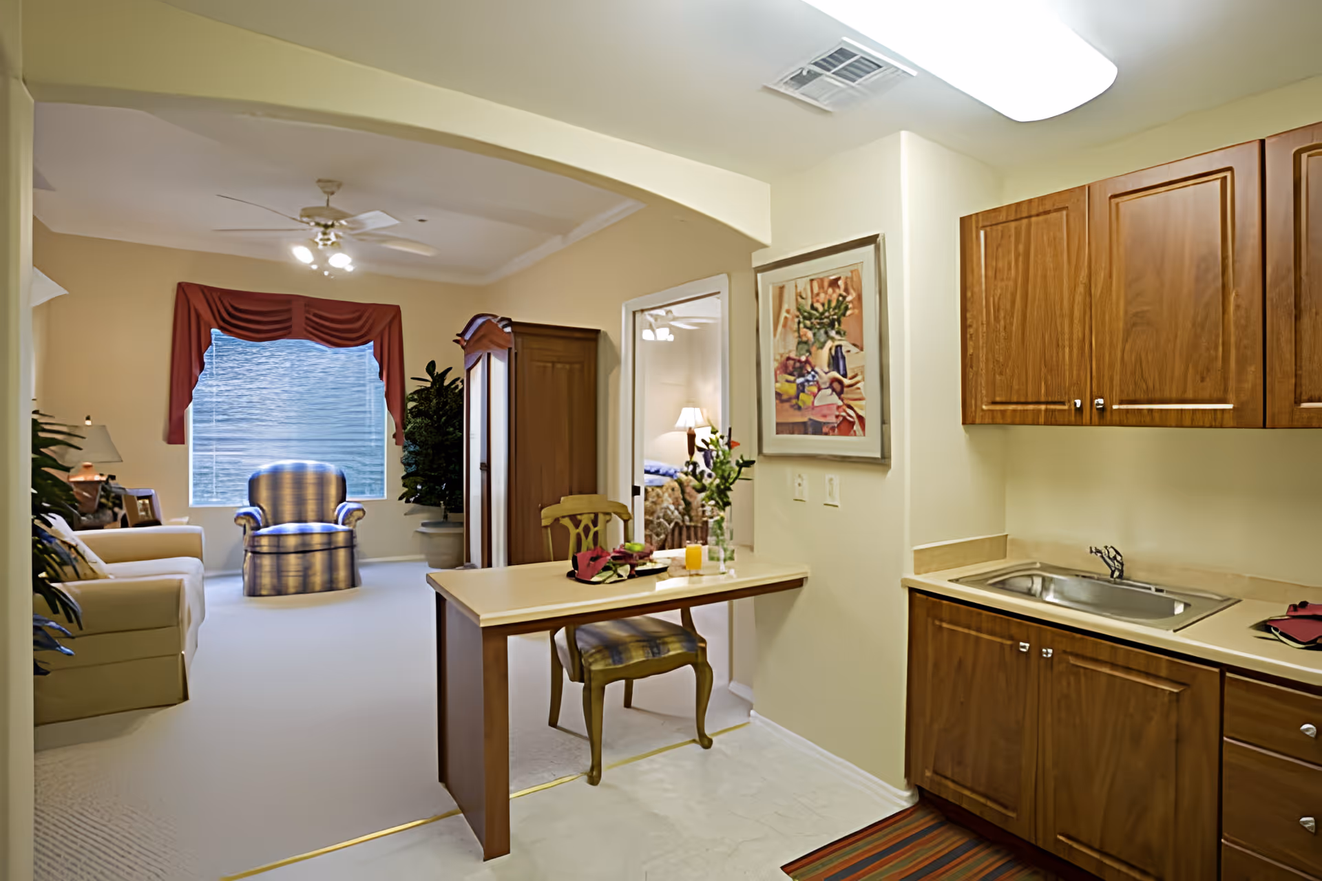 Interior view of a senior living facility showing a small kitchenette with wooden cabinets and a sink on the right, a table with a chair in the center, and a living area with armchairs, a sofa, plants, and a window with red curtains in the background. A doorway leads to a bedroom with a bed and a lamp visible.