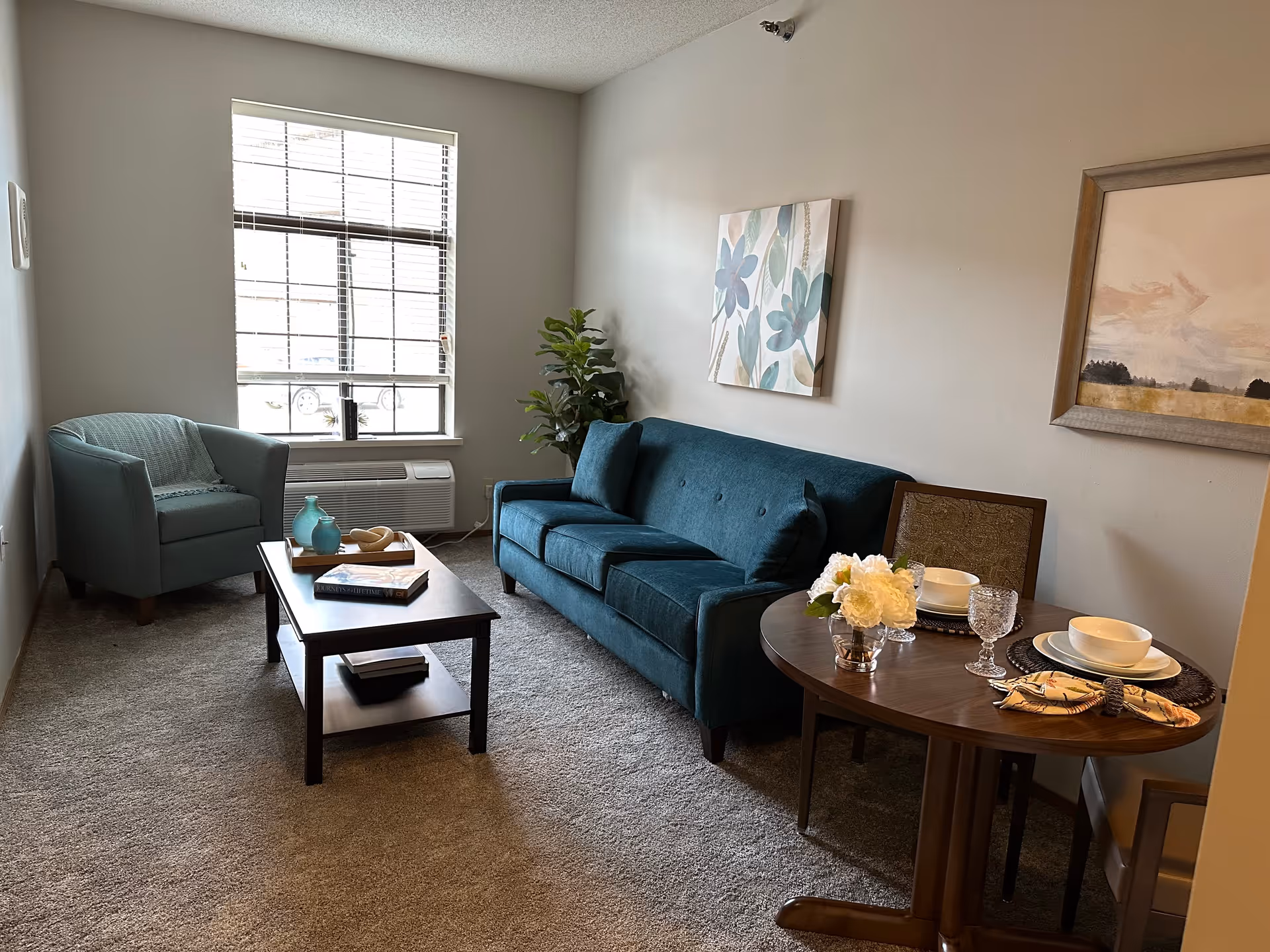 A cozy living room with a teal sofa and matching armchair, a wooden coffee table with decorative items, a small round dining table set for two with white dishes and glassware, a window with blinds, and two framed paintings on the walls.