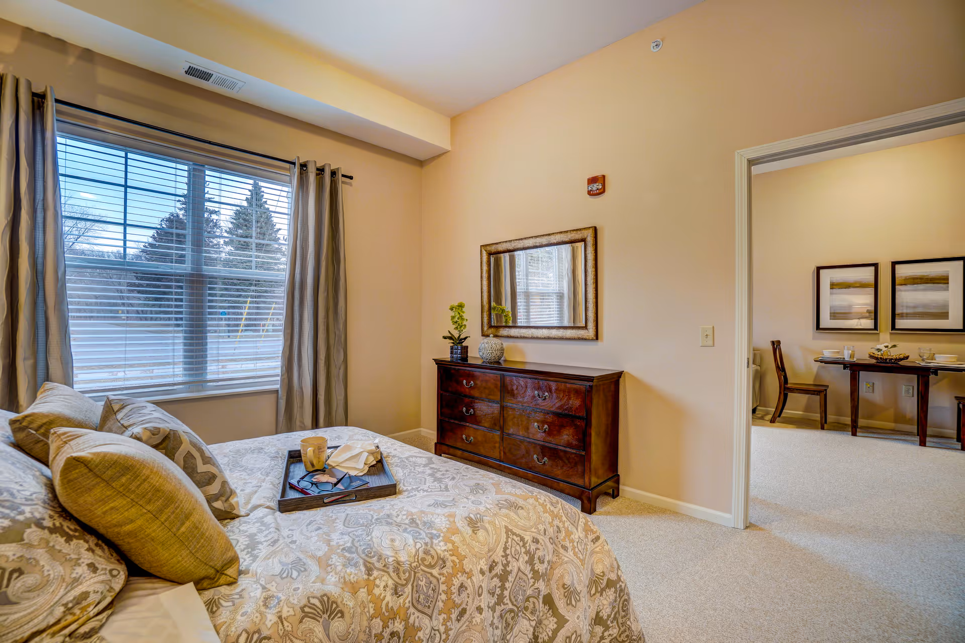 A cozy bedroom with a large window covered by curtains, a bed with patterned bedding and multiple pillows, and a wooden dresser with a mirror above it. Through an open doorway, a dining area with a table, chairs, and framed pictures on the wall is visible.