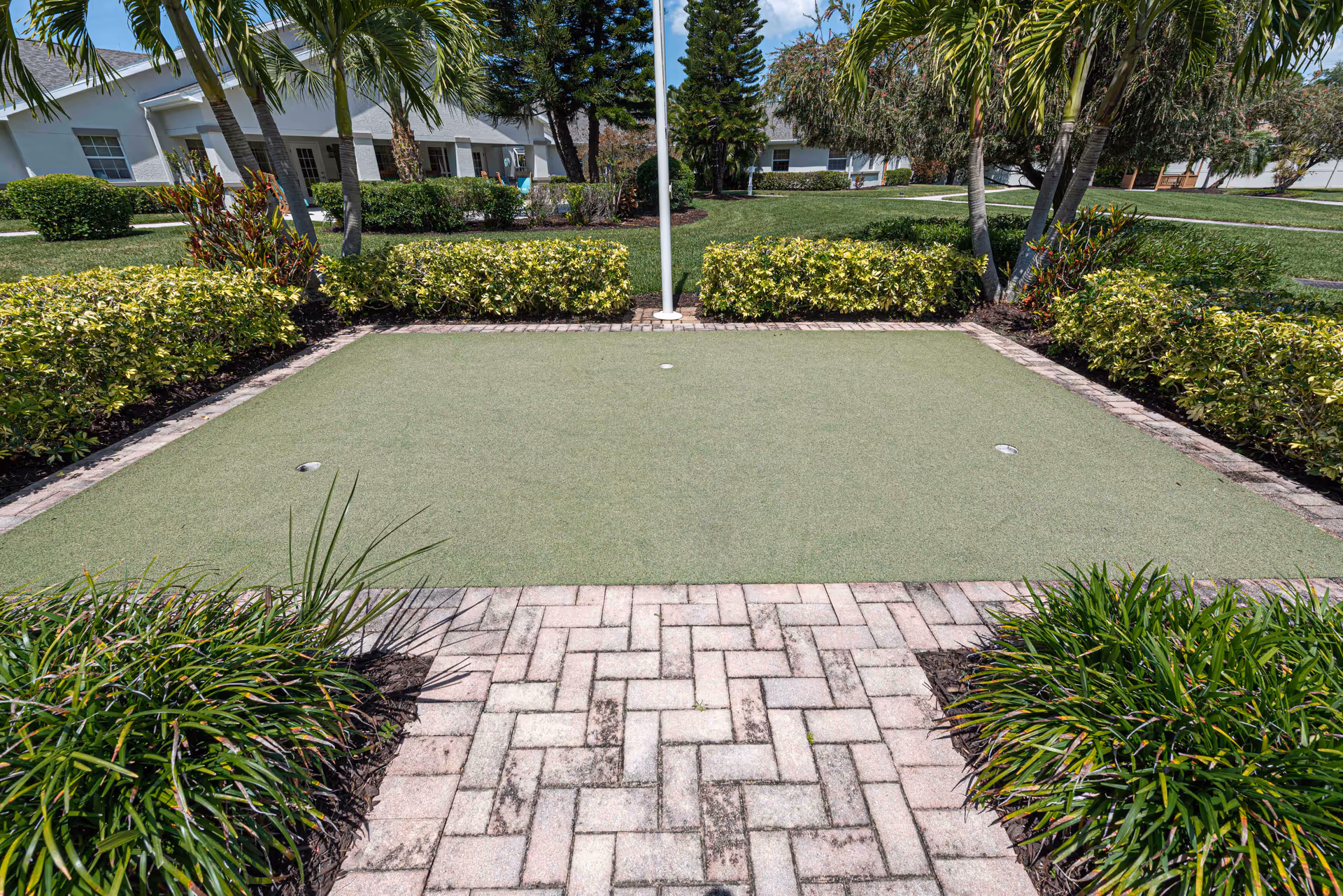 Outdoor bocce ball court surrounded by neatly trimmed bushes and palm trees, with a paved walkway leading up to it. In the background, there are white residential buildings with windows and a clear blue sky above.