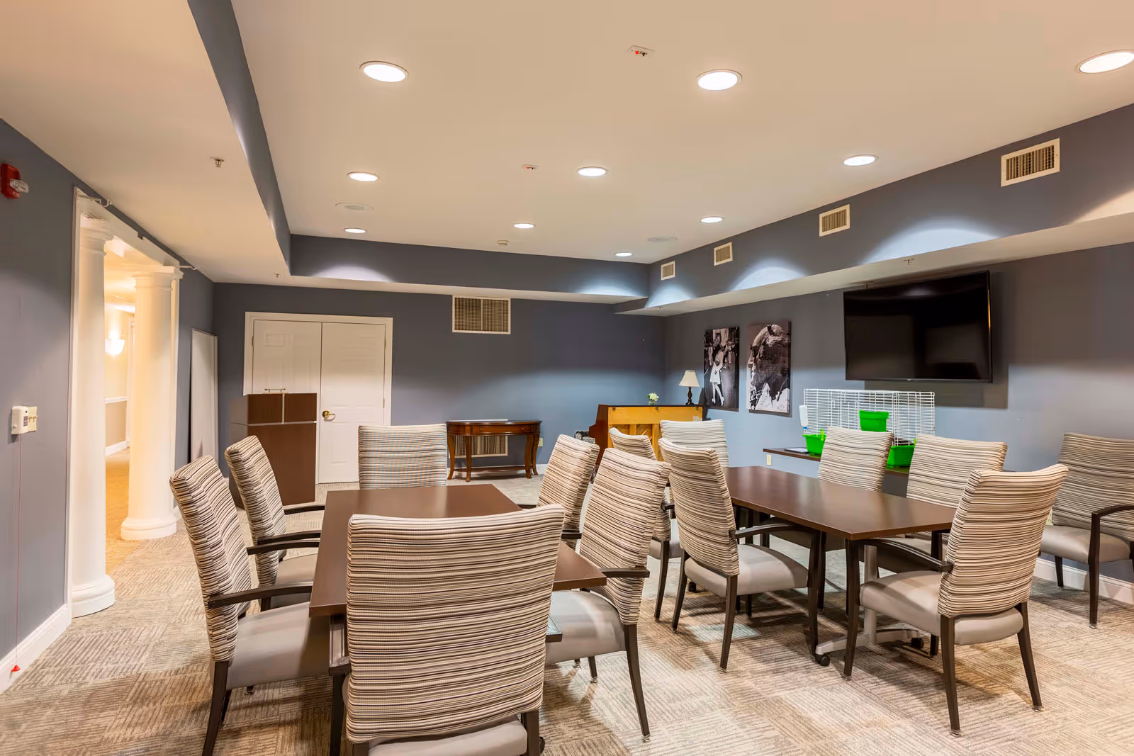 Communal dining/activity room with multiple tables and striped upholstered chairs, a wall-mounted TV, and gray-blue walls.