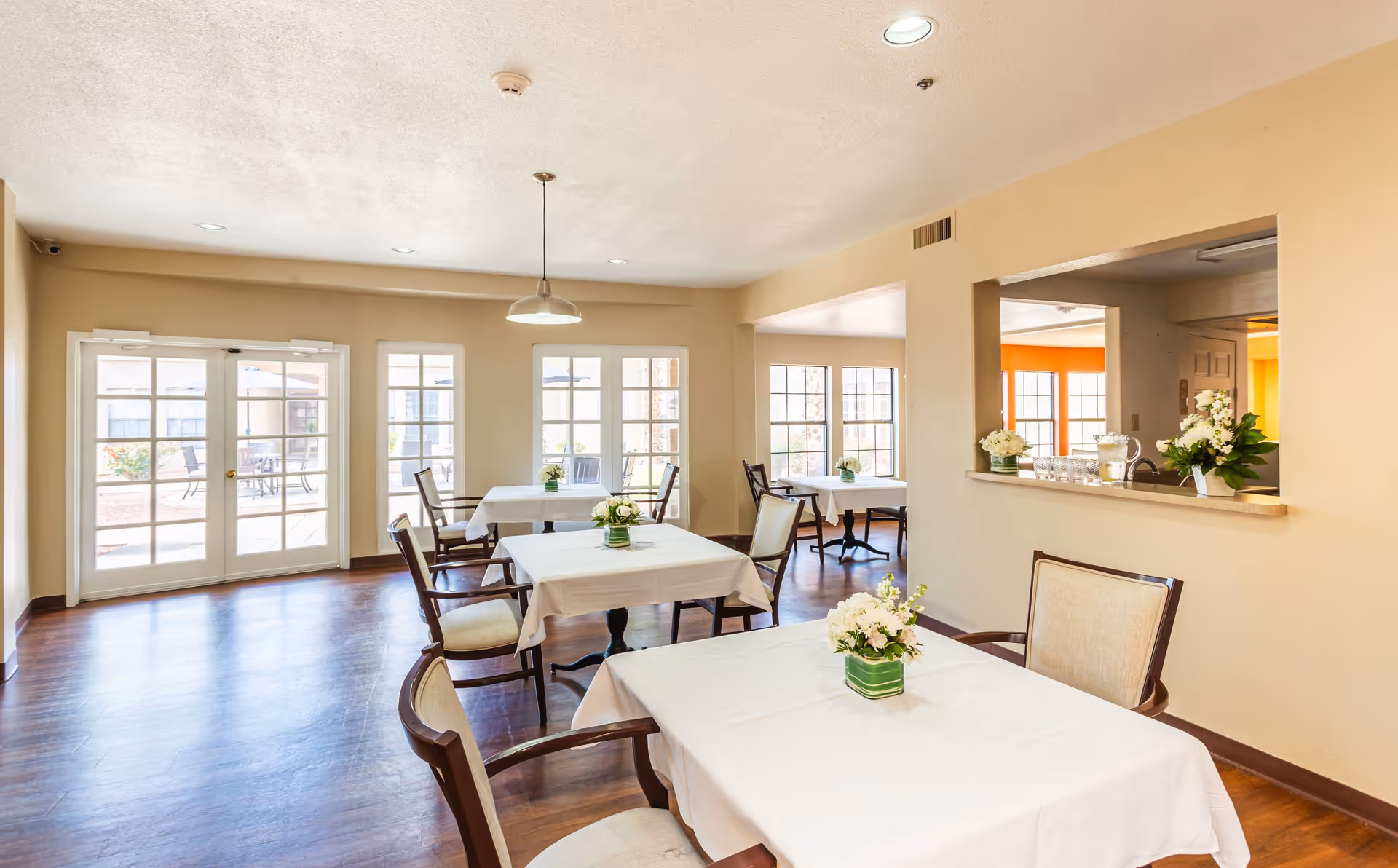 A bright dining room with several tables covered in white tablecloths, each adorned with a small floral centerpiece. The room has large windows and glass doors letting in natural light, wooden floors, and beige walls. There is a pass-through window to an adjacent room with more flowers and glassware on the counter.