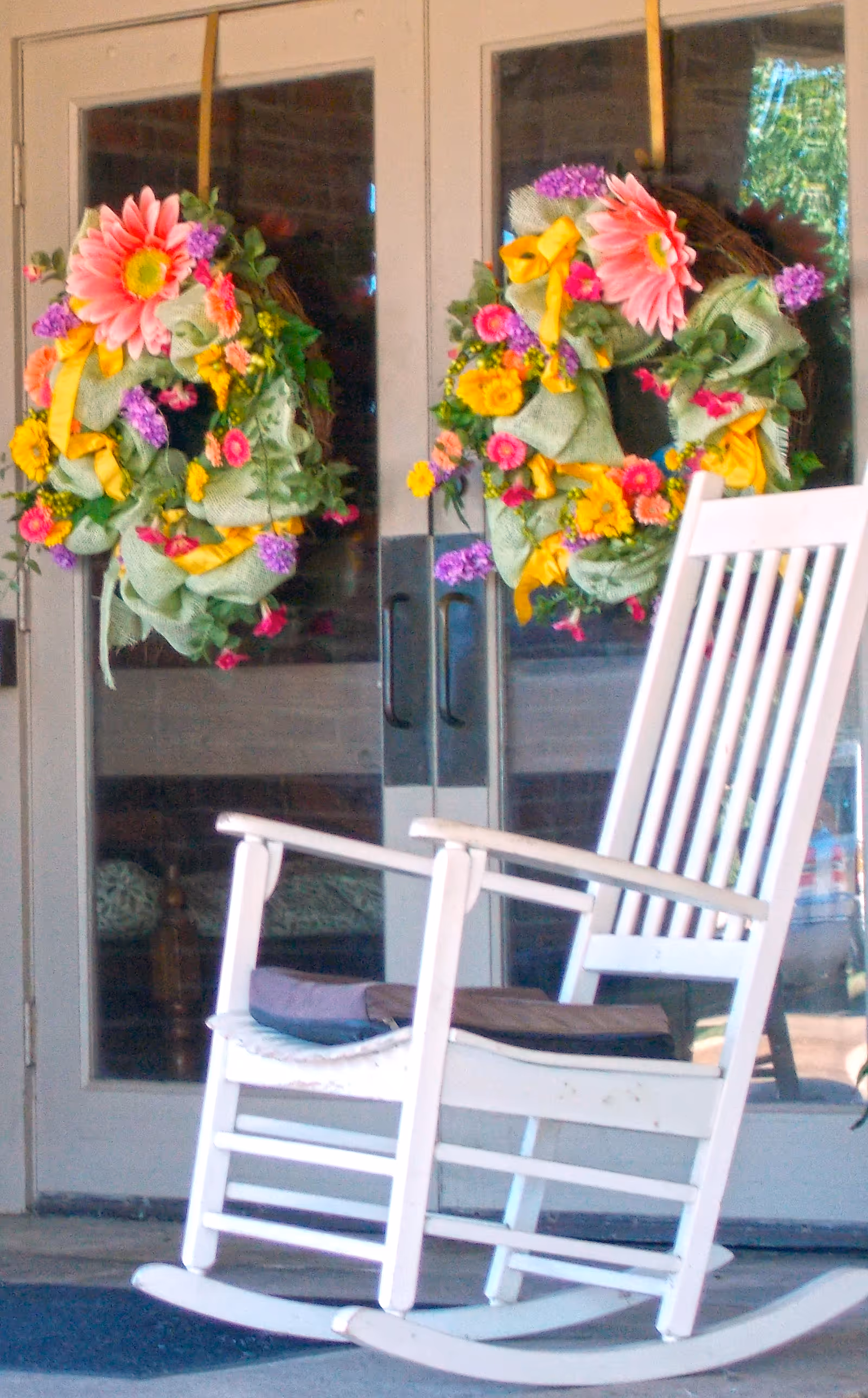 A white wooden rocking chair with a cushion placed on a porch in front of glass double doors decorated with colorful floral wreaths featuring pink, yellow, and purple flowers and green leaves.