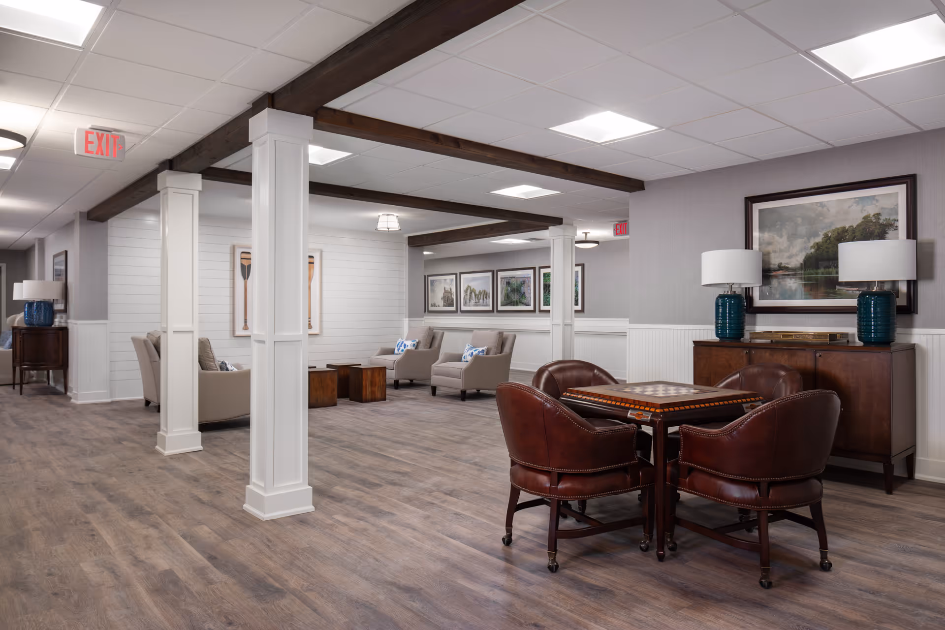 Bright communal lounge with a game table surrounded by leather chairs, upholstered seating, artwork, and decorative columns.