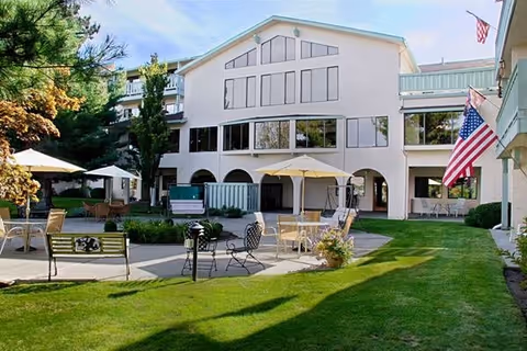 Outdoor courtyard area of a senior living facility with green grass, patio seating including tables with umbrellas, chairs, and benches. The building in the background is white with multiple windows and an American flag displayed on the right side.