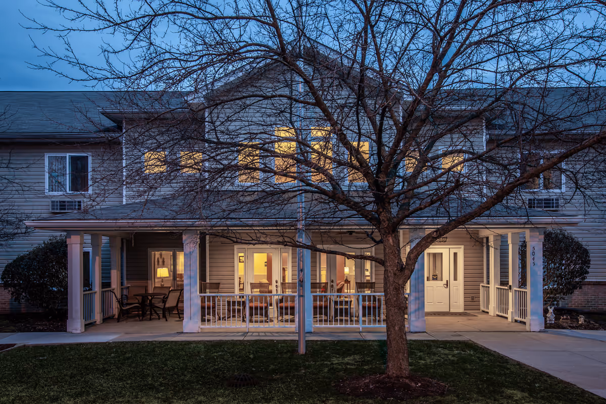 Exterior view of a two-story senior living facility at dusk with warm lights glowing from the windows. The building has a covered porch with seating and white railings. A large leafless tree stands in front of the building on a grassy lawn.
