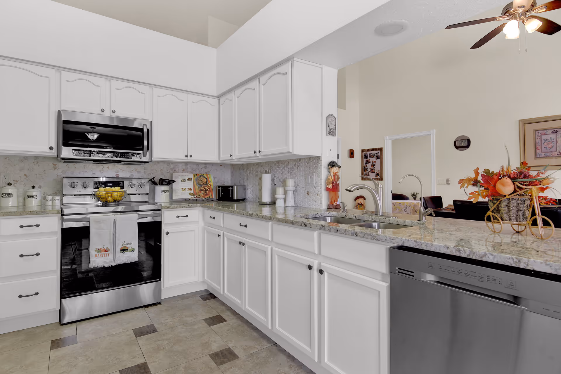 Bright and modern kitchen with white cabinets, granite countertops, stainless steel appliances including a stove, microwave, and dishwasher. The kitchen has a tiled floor and a ceiling fan with lights. There is a decorative basket with autumn-themed flowers on the counter and a view into a living area with a clock and framed pictures on the wall.