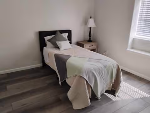 A simple bedroom with a single bed covered with a beige blanket and a gray throw. There are two pillows on the bed, one gray and one white. Next to the bed is a wooden nightstand with a black lamp. Sunlight streams in through a window with white blinds, casting shadows on the floor.