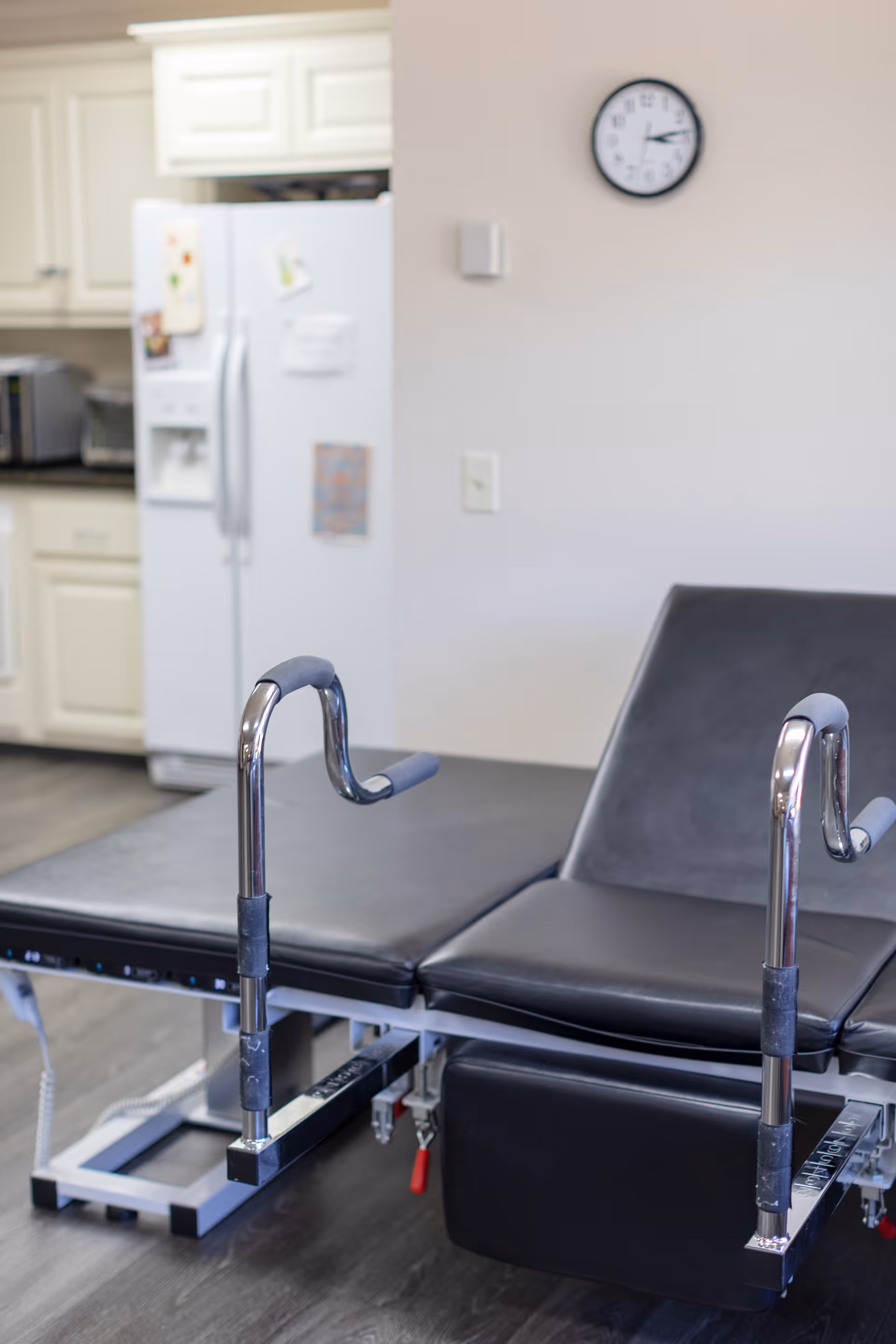 A medical examination table with black padding and metal stirrups in a room with a white refrigerator, kitchen cabinets, and a wall clock showing 2:15.