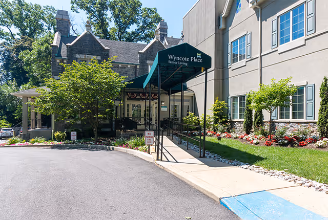 Entrance to Wyncote Place senior living facility with a covered walkway, landscaped garden with flowers and shrubs, and a paved driveway. The building has stone and beige siding with multiple windows.