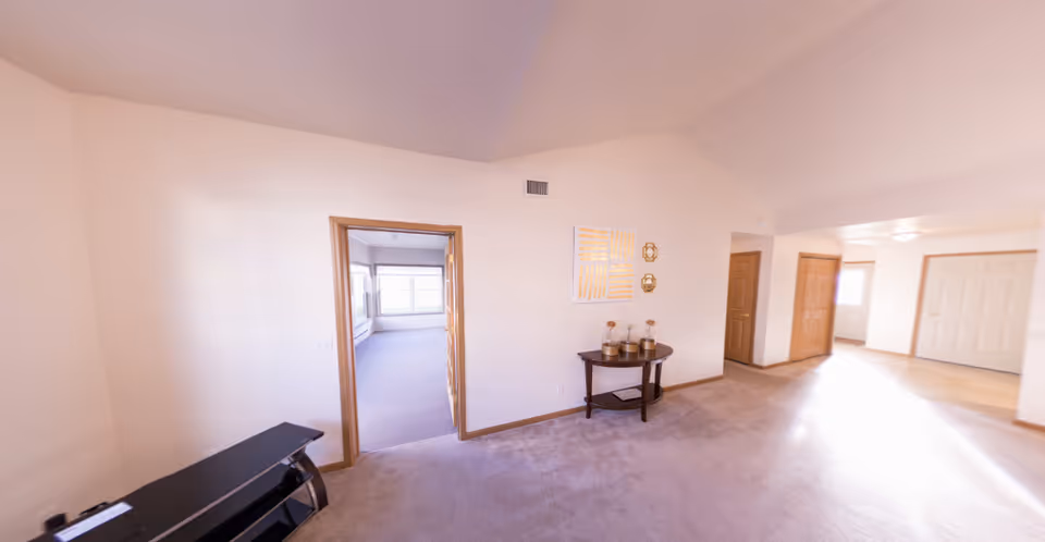 Spacious carpeted living area with a small console table and decorative items, a doorway to an adjacent sunlit room, and an entry hallway.