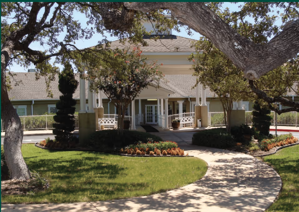 Front entrance of Independence Hill Retirement Community with a curved walkway leading to a covered porch. The area is landscaped with green grass, trees, shrubs, and flower beds under a clear sky.