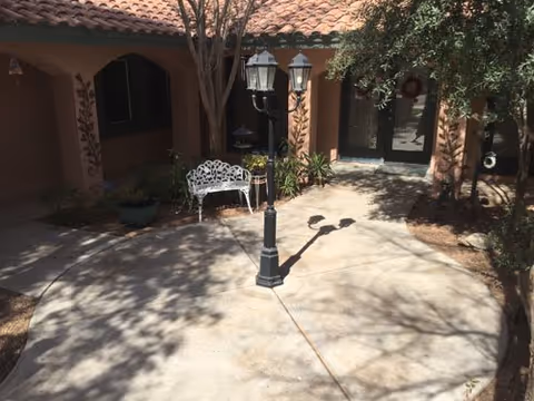 Outdoor courtyard area with a concrete circular patio, a black vintage-style street lamp in the center, a white metal bench, potted plants, and trees casting shadows. The background shows a building with a tiled roof and arched doorways.