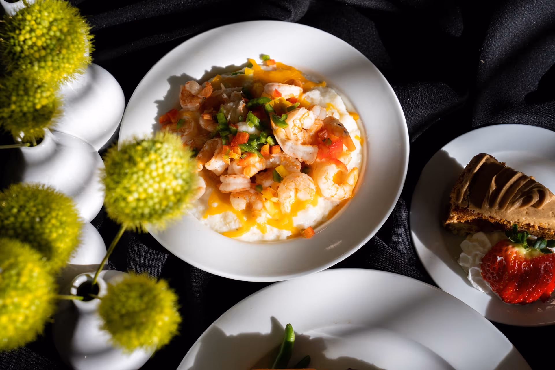 A white plate with shrimp served over a bed of creamy grits topped with diced green and red bell peppers. Next to it is a smaller white plate with a slice of dessert garnished with a strawberry and whipped cream. Green spherical flowers in white vases are partially visible on the left side, all set on a black tablecloth.