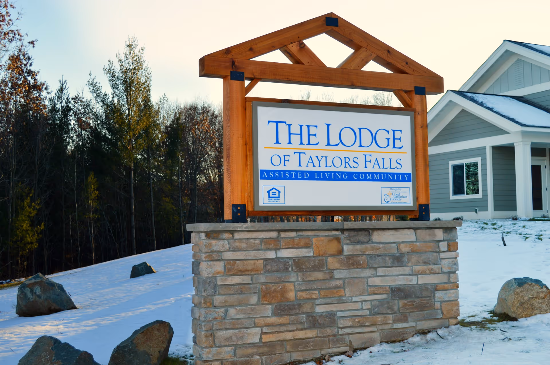 A wooden and stone sign for The Lodge of Taylors Falls Assisted Living Community stands outdoors on a snowy ground with trees in the background and a gray building with white trim partially visible to the right.