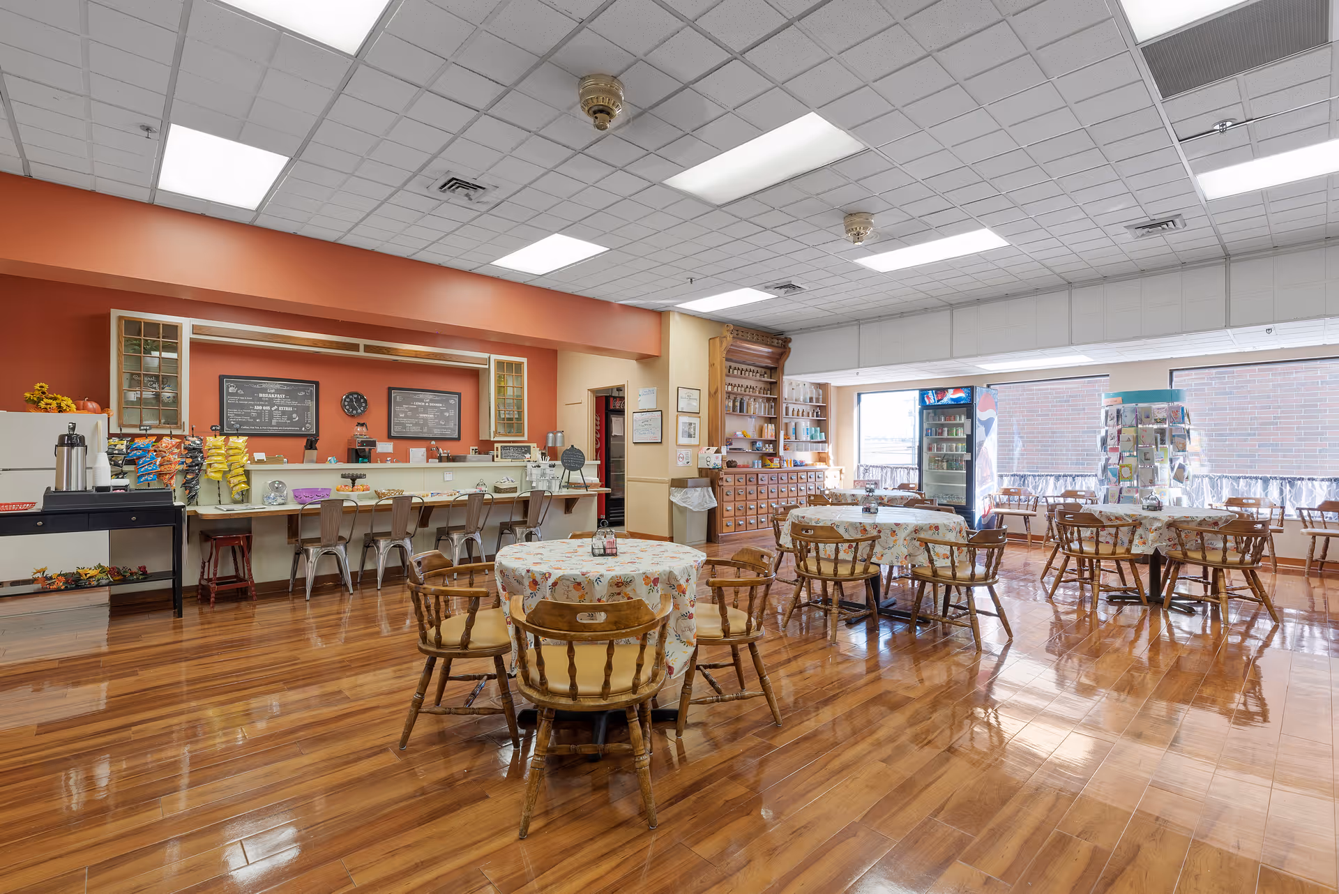 A bright and spacious dining area in Cottingham Retirement Community featuring several round tables covered with floral tablecloths and surrounded by wooden chairs. The room has polished wooden floors, a counter with stools along one wall, and large windows letting in natural light. There is a beverage cooler and a display rack near the windows, and the walls are painted in warm tones with some decorative elements.