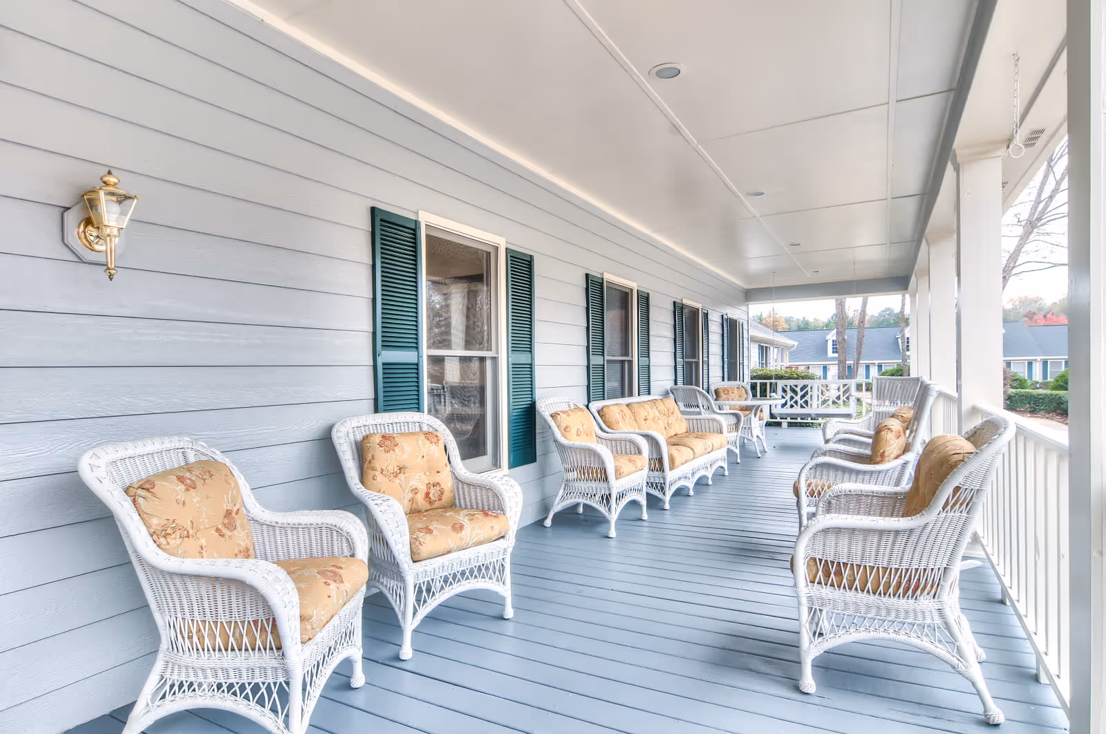 A spacious covered porch with white wicker chairs and sofas featuring floral cushions, light blue wooden floorboards, light blue siding with green shutters on windows, and white ceiling with recessed lighting. The porch overlooks a landscaped outdoor area with trees and shrubs.