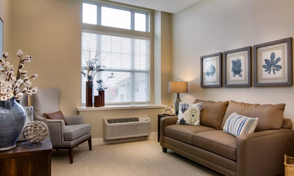 Bright living room with a brown sofa, upholstered armchair, window with blinds, side table and framed botanical artwork.