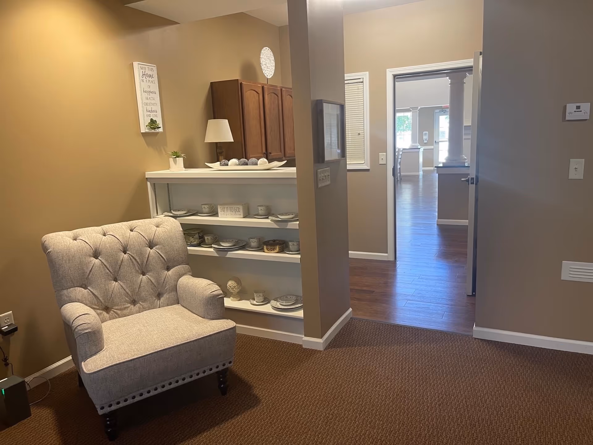 A cozy interior corner of a senior living facility with a beige tufted armchair on a brown carpet. Behind the chair is a white shelving unit displaying decorative plates, teacups, and a small sign. The walls are painted light brown, and there is a doorway leading to a room with wooden flooring and white columns visible in the distance.