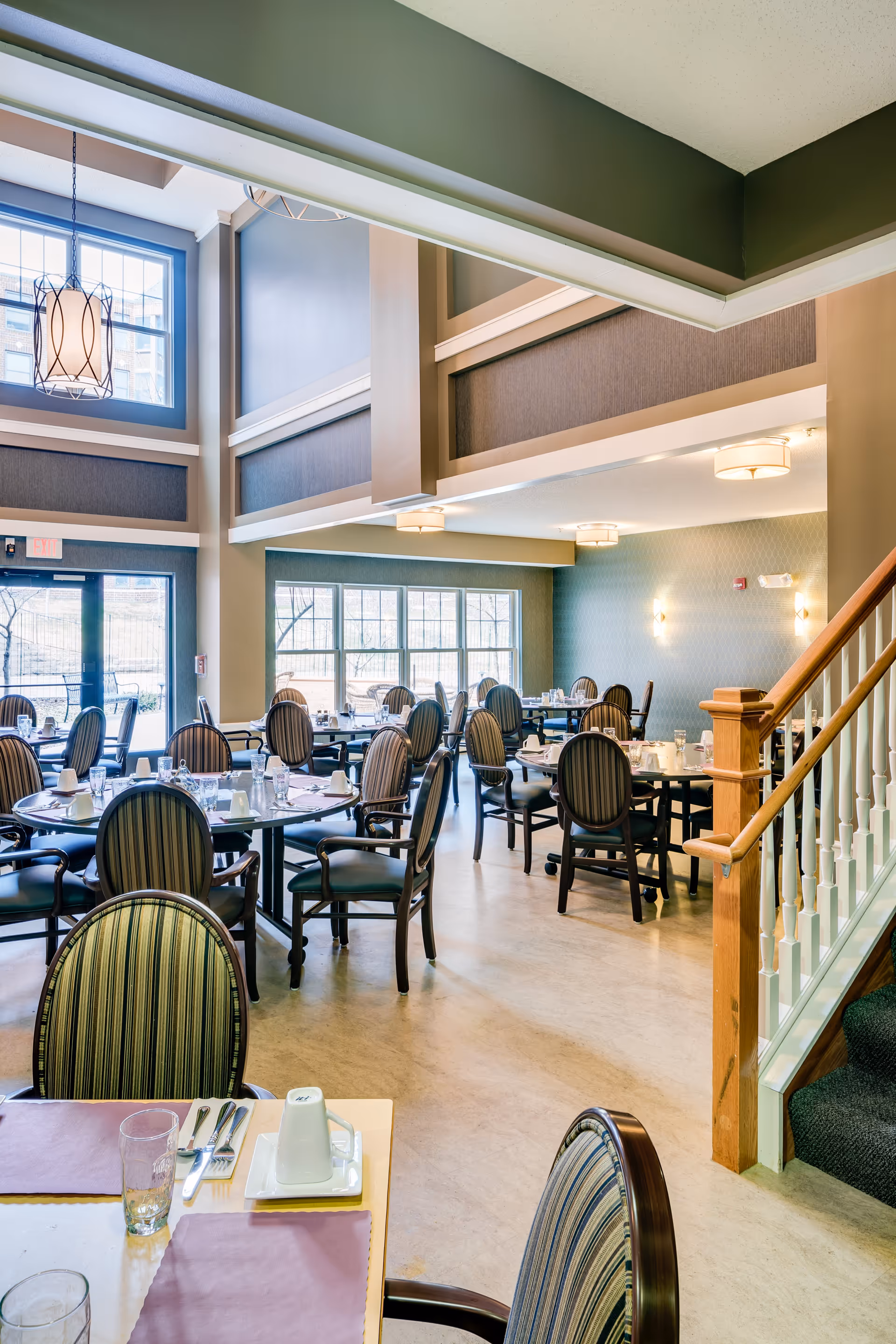 Bright, spacious dining room with multiple set tables and striped chairs near a staircase and large windows.