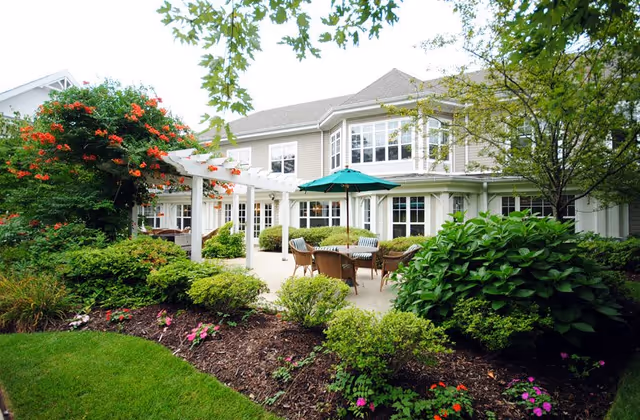 Outdoor patio area at Sunrise of Holbrook featuring a seating arrangement with wicker chairs and a table under a green umbrella, surrounded by lush greenery, flowering plants, and a white pergola attached to a two-story building with multiple windows.