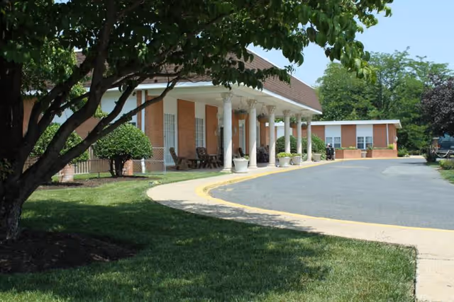 Exterior view of a brick senior living facility with a covered entrance supported by white columns, a curved driveway, and landscaped grounds.