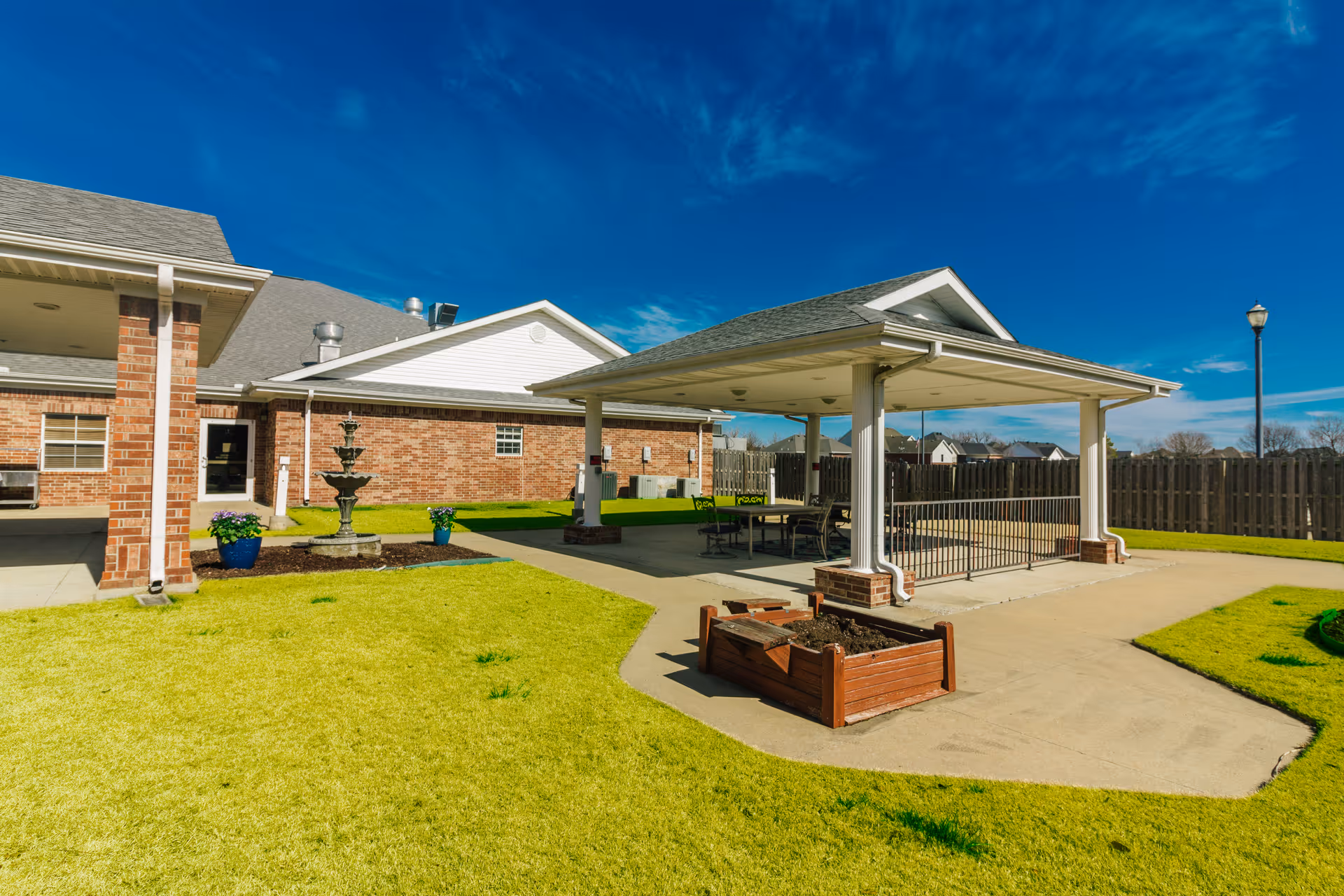 Outdoor patio area at The Maples at Har-Ber Meadows featuring a covered seating area with tables and chairs, a brick building in the background, a small fountain, potted plants, and a raised garden bed on a sunny day with a clear blue sky.