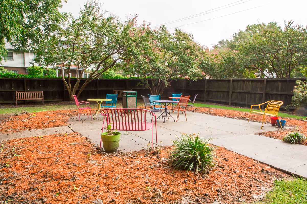Outdoor courtyard with colorful metal chairs and tables on a concrete patio surrounded by mulch beds, trees, and a wooden fence.