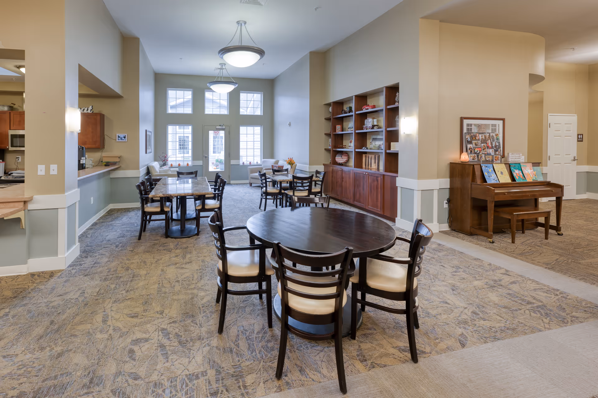 A spacious common area with multiple round and rectangular tables surrounded by chairs. The room features large windows letting in natural light, a built-in wooden shelving unit with decorative items, and a piano with colorful artwork displayed on top. The walls are painted beige with white trim, and the carpet has a patterned design.