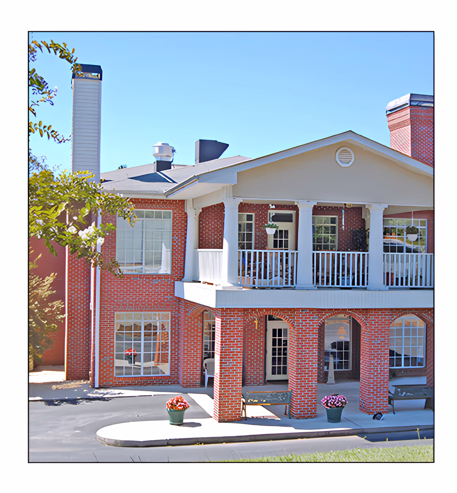 Exterior view of a two-story brick building with white columns and railings on the upper balcony. There are large windows on both floors, flower pots with red and pink flowers near the entrance, and benches under the covered area. The sky is clear and blue.
