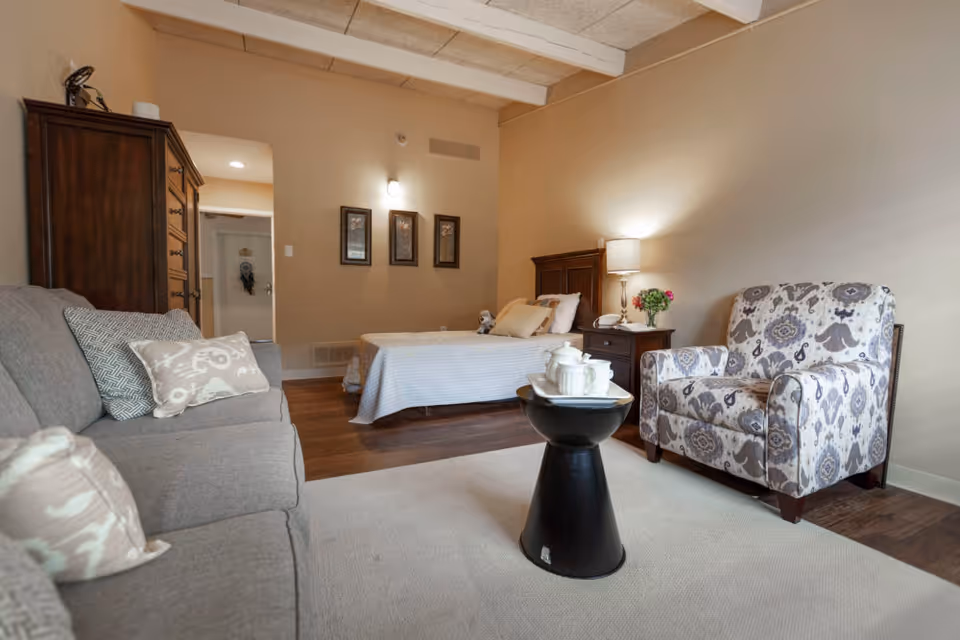 A cozy room with a bed in the background, a wooden nightstand with a lamp and flowers, a patterned armchair, a small black side table with a white tea set, and a gray sofa with decorative pillows in the foreground. The walls are beige and there are three framed pictures on the wall near the bed.
