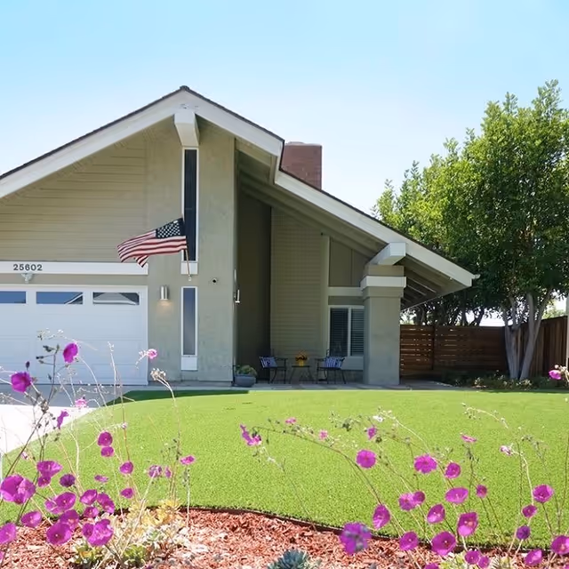 Front exterior view of a single-story house with a slanted roof, an American flag mounted near the entrance, a garage door on the left, and a small patio area with two chairs and a table. The lawn is green and well-maintained with purple flowers in the foreground and trees on the right side.