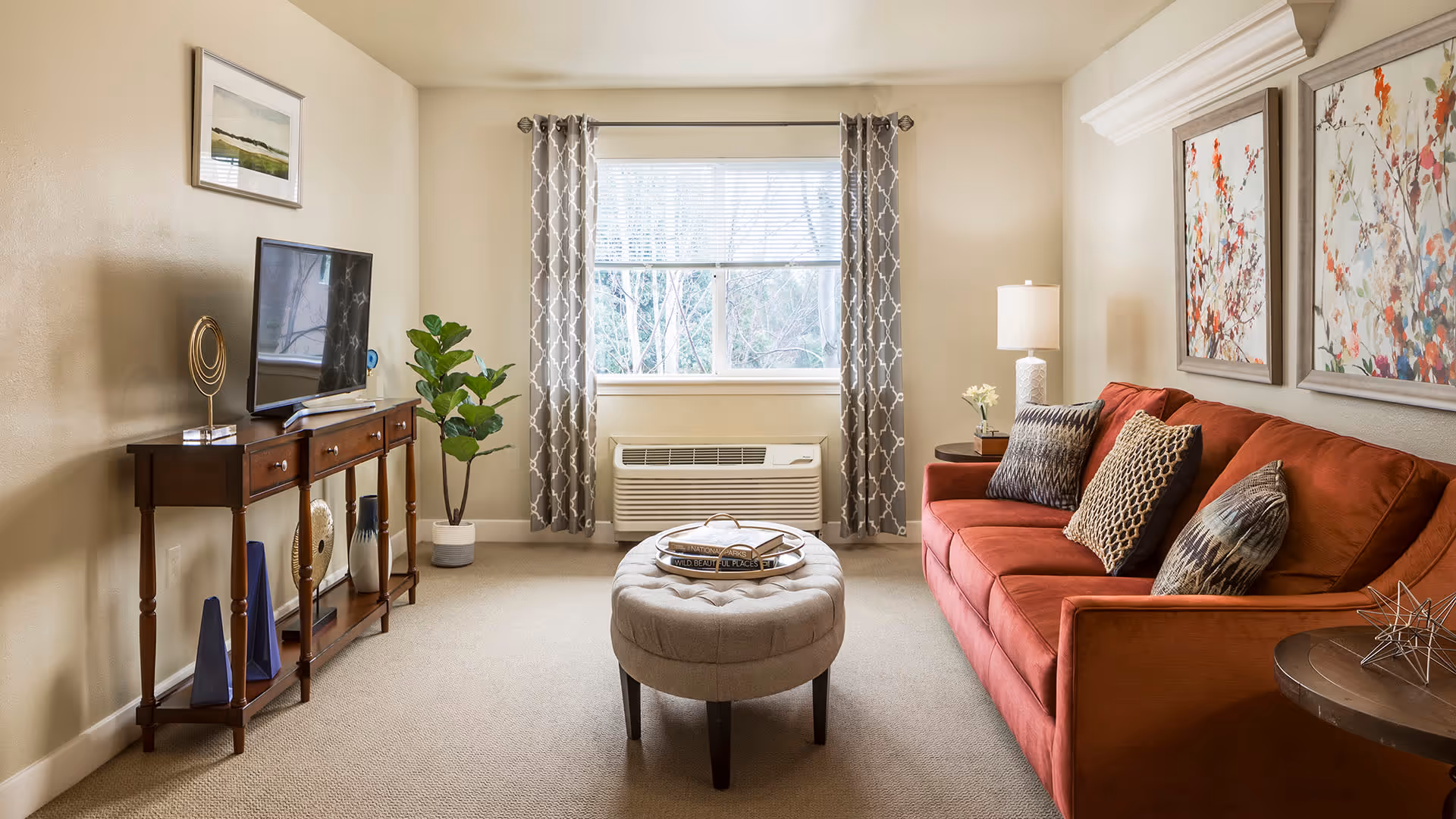 A cozy living room with a red sofa adorned with patterned cushions, a round tufted ottoman in the center with books on top, a wooden console table holding a flat-screen TV and decorative items, a potted plant beside the window with patterned curtains, and two framed floral artworks on the wall.