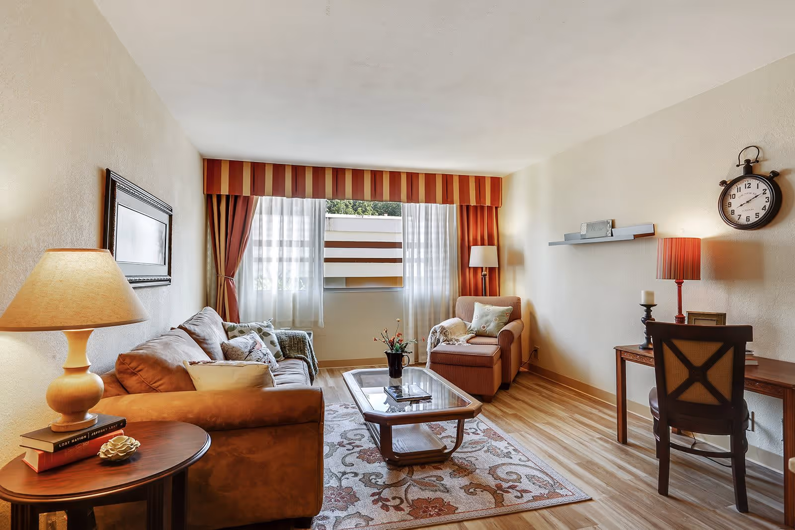 A cozy living room with a brown sofa and armchair, a wooden coffee table on a patterned rug, a side table with a lamp and books, a desk with a chair and lamp, a wall clock, and a window with striped valance and curtains letting in natural light.