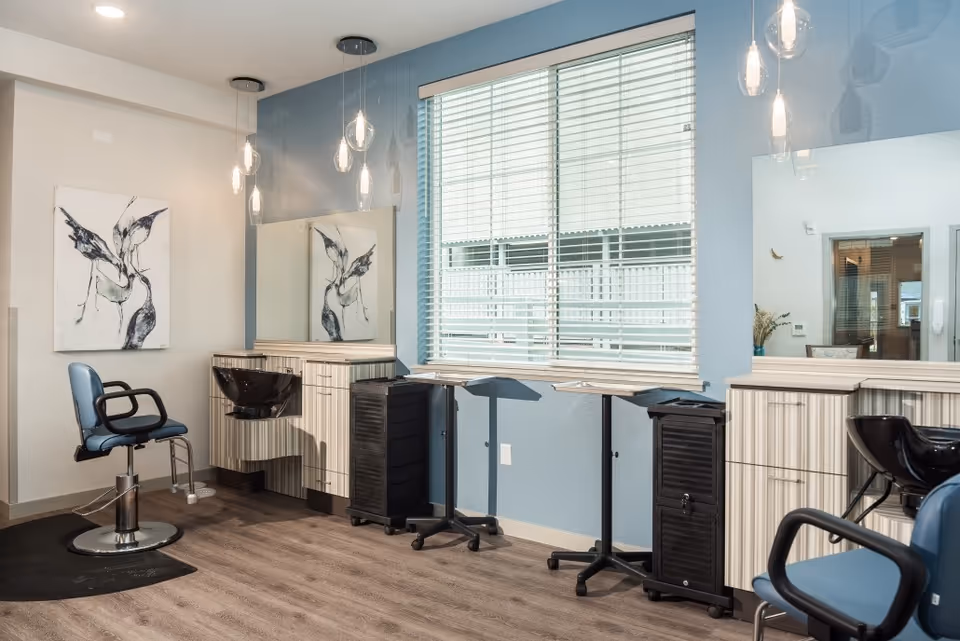 A bright salon with styling chairs, wash basins, mirrors, and a large window with blinds.
