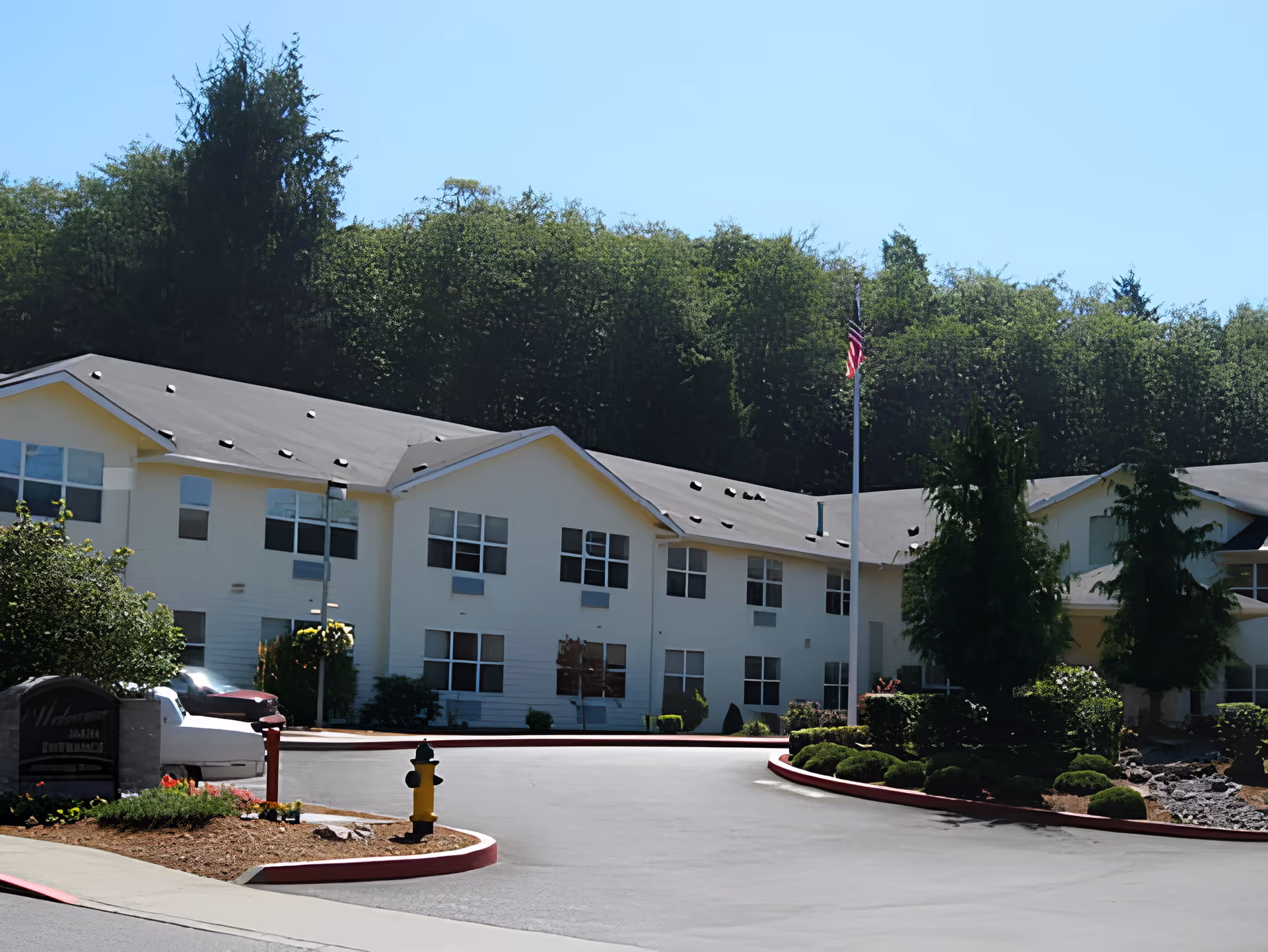 Exterior view of a two-story assisted living facility building with multiple windows, surrounded by trees and landscaped bushes. There is a driveway with a yellow fire hydrant and an American flag on a flagpole near the entrance.