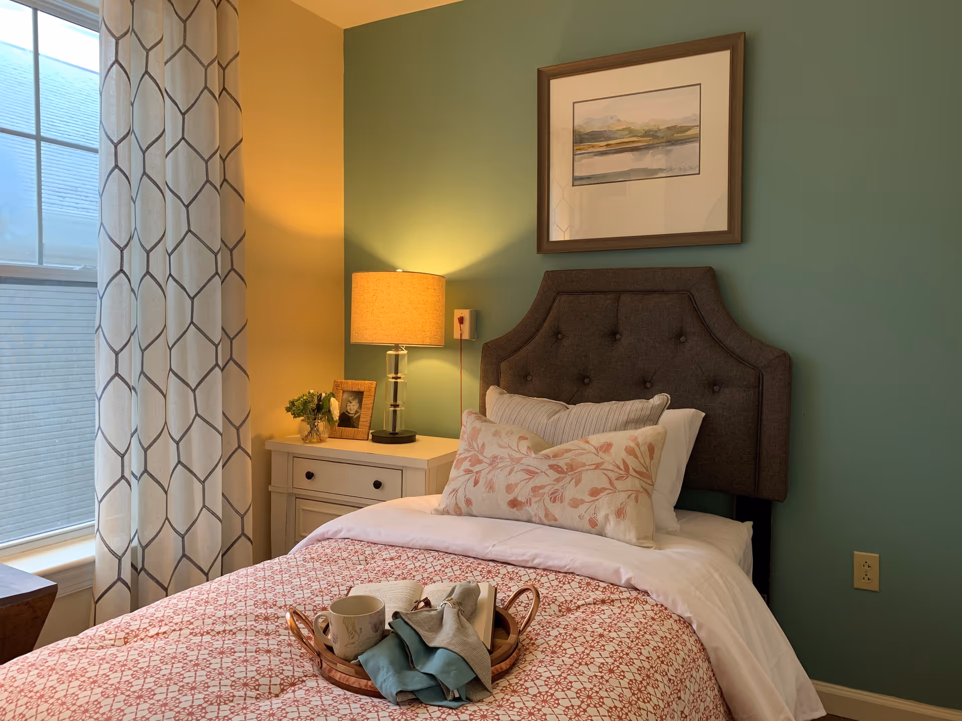 A cozy bedroom with a single bed featuring a dark tufted headboard, white and floral pillows, and a red patterned quilt. A wooden tray with a cup, an open book, and a cloth is placed on the bed. Next to the bed is a white nightstand with a table lamp, a small plant, and a framed photo. The walls are painted green and yellow, and a window with patterned curtains lets in natural light.