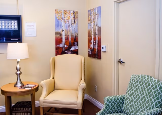 A cozy corner of a senior living community room featuring a beige upholstered armchair, a green patterned armchair, a round wooden side table with a lamp and a magazine, and two colorful paintings of birch trees on the wall.