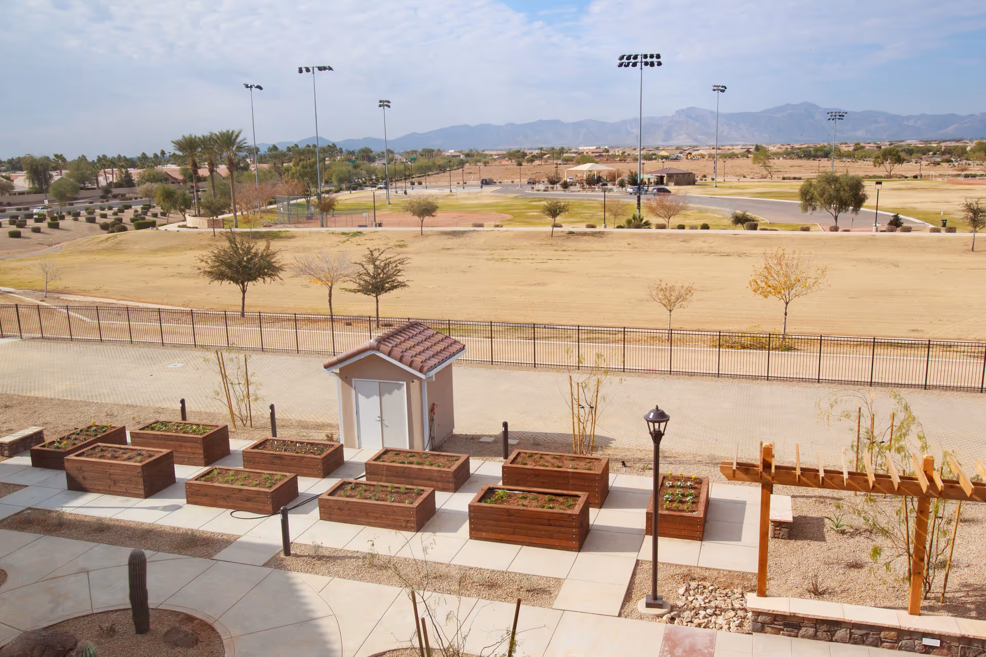 View of an outdoor garden area with multiple wooden raised garden beds containing small plants. There is a small shed with a tiled roof, a lamppost, and a wooden pergola. Beyond the garden area, there is a fenced open field with scattered trees, sports field lighting, and mountains in the background under a partly cloudy sky.