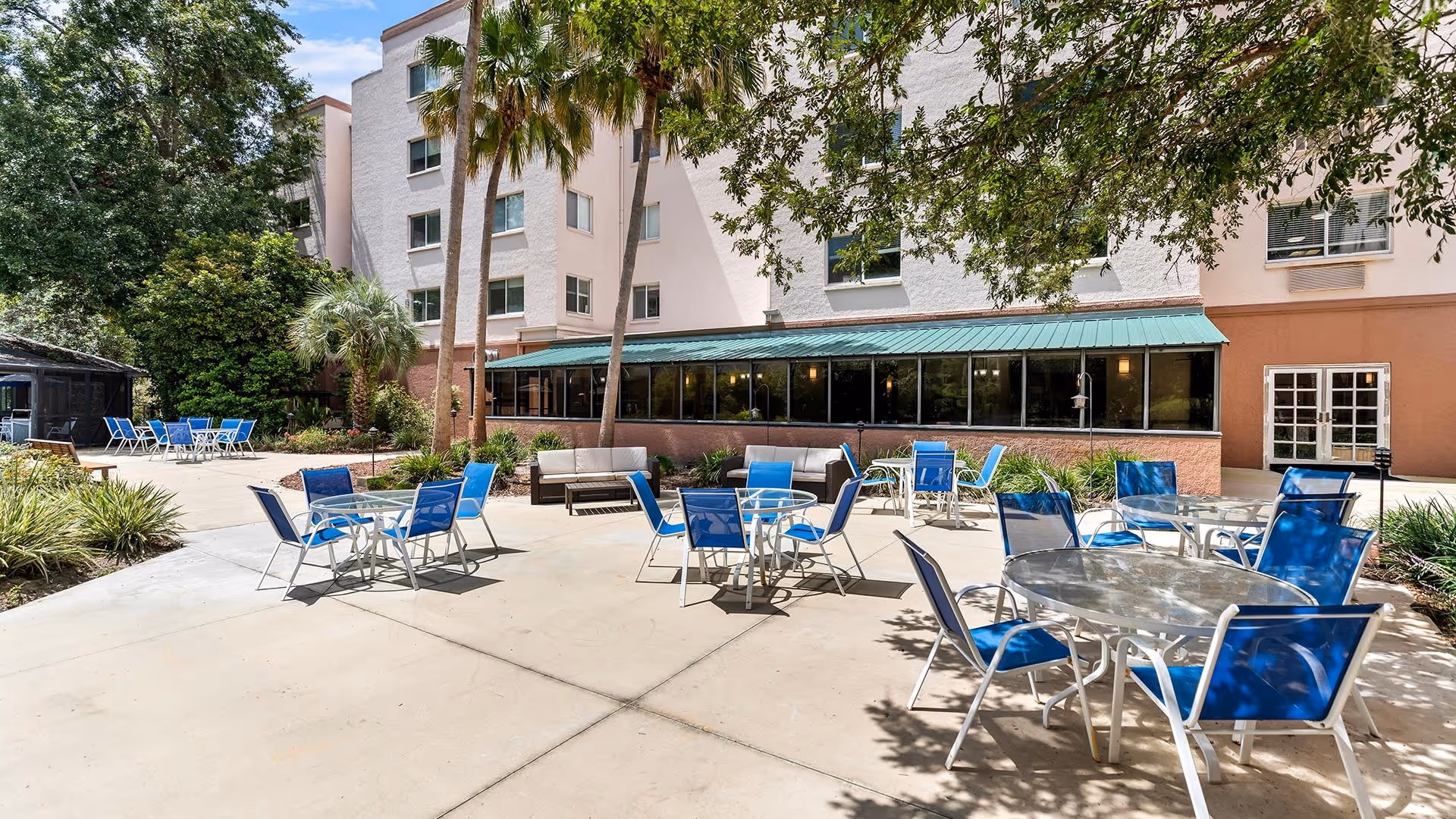 Outdoor patio area at Atrium at Gainesville with multiple round glass tables and blue chairs arranged on a concrete surface. There are some cushioned seating areas along the building wall. The building has multiple floors with windows, and there are palm trees and other greenery surrounding the patio.