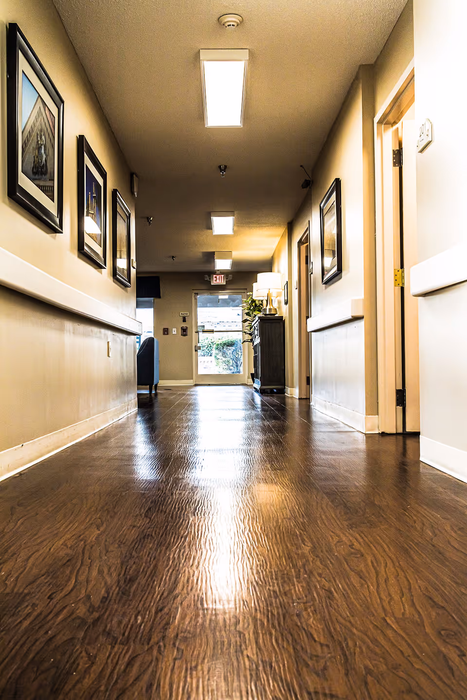 A well-lit hallway with wooden flooring in a senior living facility. The walls are adorned with framed pictures, and there is a cabinet with a lamp and a plant near the end of the hallway. An exit door with a glass window is visible at the far end.