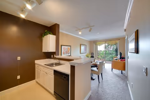 Open-plan apartment interior with a small kitchen and breakfast bar leading to a living and dining area with a sliding glass door to a balcony.