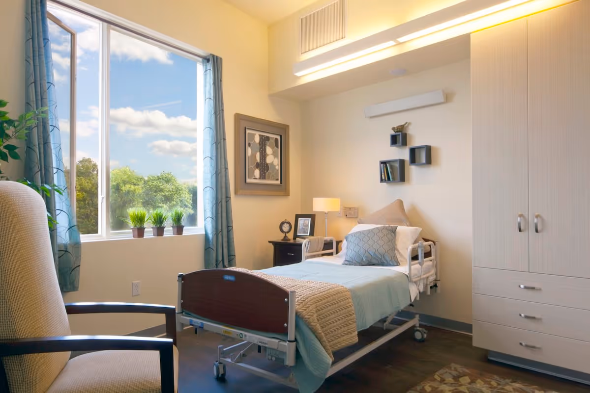 A bright and cozy room in a care facility featuring a single hospital-style bed with light blue and beige bedding, a beige armchair, a large window with blue curtains showing a view of trees and sky, a small black nightstand with a lamp, clock, and picture frame, wall-mounted shelves with books and a decorative bird, and a light wood wardrobe with drawers.