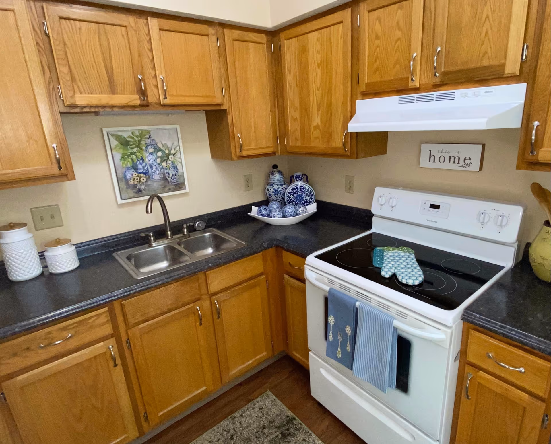 Compact kitchen with oak cabinets, black countertops, a double sink, and a white electric stove with decorative items.