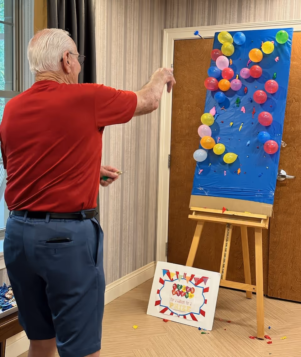 An elderly man wearing a red shirt and blue shorts is playing a balloon dart game indoors. He is throwing a dart at a board covered with colorful balloons mounted on an easel. A sign on the floor next to the easel reads 'Pop 3 balloons for a prize.' The room has beige striped wallpaper and a wooden door in the background.