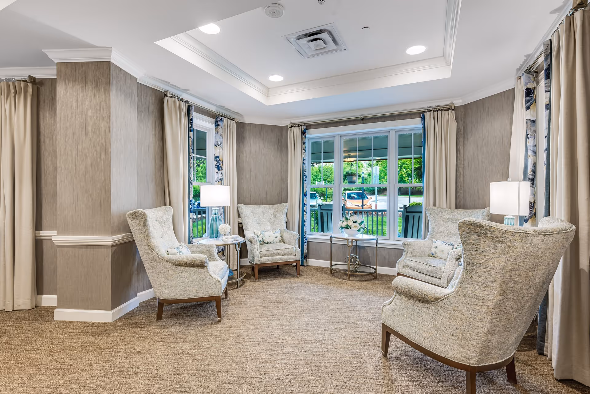 A cozy sitting area with four upholstered armchairs arranged around two small round tables, each with a lamp. Large windows with beige curtains and floral drapes let in natural light, showing a view of greenery and parked cars outside. The room has beige carpet, light brown walls, and a tray ceiling with recessed lighting.