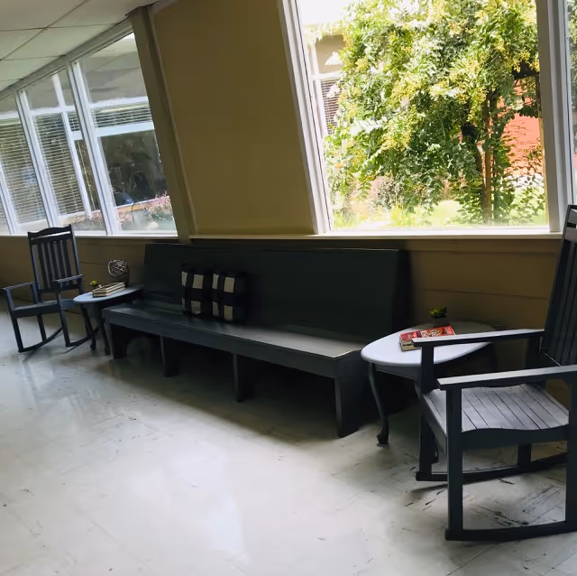 A bright indoor seating area with large windows showing green trees outside. The seating includes a long dark bench with two checkered pillows, two wooden chairs, and two small round tables with books and a small plant on them. The floor is light-colored and shiny.