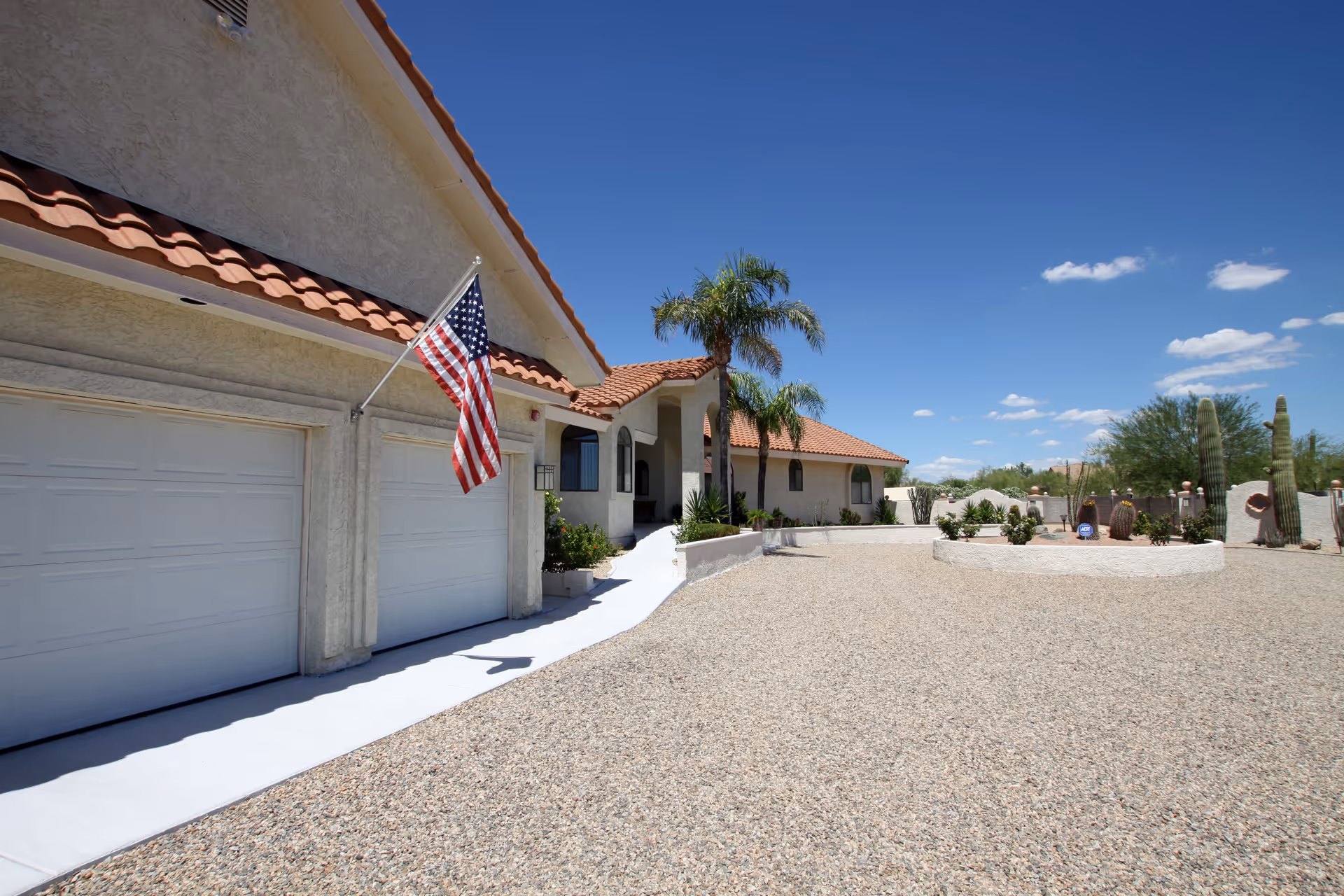 Exterior view of a single-story building with a tiled roof and beige stucco walls. There is an American flag mounted near two closed garage doors. The front yard is covered with gravel and features desert landscaping with cacti and palm trees under a clear blue sky.