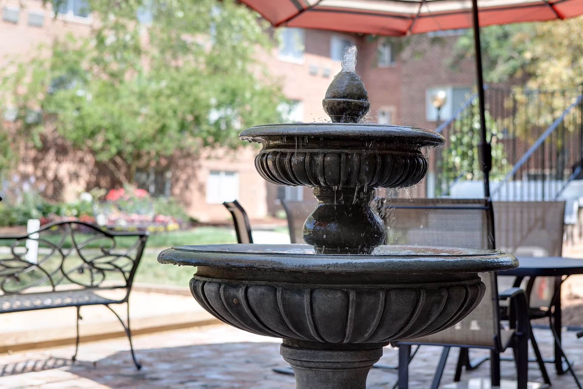 Stone tiered fountain in a courtyard with patio chairs, tables and a brick building in the background.