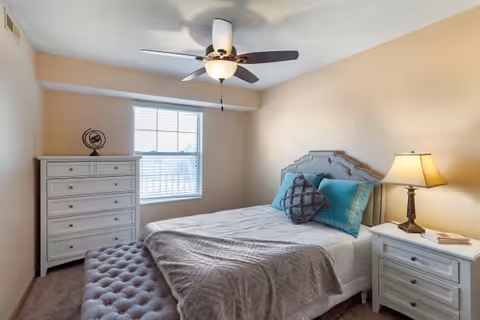 A cozy bedroom with a bed featuring a gray headboard, blue and gray pillows, and a gray blanket. There is a white nightstand with a lamp and a book on the right side of the bed. A white dresser with multiple drawers is positioned against the wall near a window with blinds. A ceiling fan with a light fixture is mounted on the ceiling. The walls are painted beige.